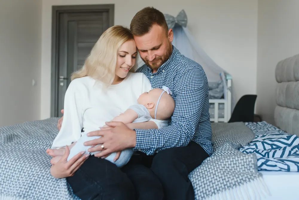 A couple sitting together on a bed at home holding their newborn baby